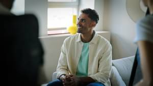 Oscar da Silva sitzt seitlich auf der Couch in einem Wohnzimmer und lächelt freundlich. Um ihn herum sind Personen des Kamera- und Interviewteams angeschnitten.