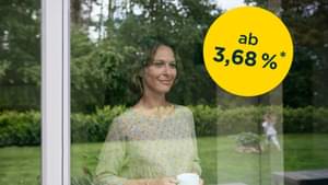 Frau steht mit ihrer Kaffeetasse am Fenster schaut ihrer Tochter beim Spielen zu: Baufinanzierung ab 3,68% p.a.*