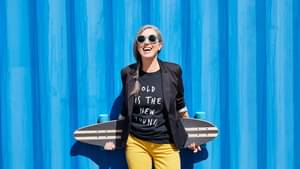 Ältere Frau steht lachend vor einer blauen Wand und hält lässig ein Longboard hinter ihrem Rücken.