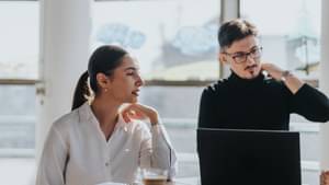 Zwei junge Berufstätige sitzen an einem Schreibtisch in einem hellen Büro. Die Frau trägt eine weiße Bluse und spricht, während der Mann im schwarzen Rollkragenpullover nachdenklich auf einen Laptop blickt.