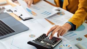 Eine junge Frau im gelben Blazer und blauem Pullover sitzt an ihrem Schreibtisch, schaut sich Bilanzen und Diagramme auf ausgedruckten Dokumenten an und tippt etwas in einen Taschenrechner ein.