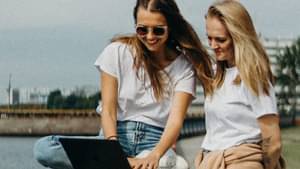 Junge Frauen, die auf einer Mauer an einem Flussufer sitzen und gemeinsam auf einen Laptop schauen.