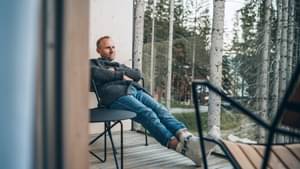 Mann mit Jeans und Wollpullover sitzt entspannt auf Balkon mit Blick auf Birkenwald.