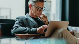 Mann sitzt nachdenklich auf einem Sessel, mit Laptop auf dem Schoß und checkt sein Aktien-Depot.