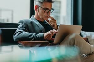 Mann sitzt nachdenklich auf einem Sessel, mit Laptop auf dem Schoß und checkt sein Aktien-Depot.