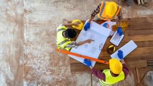 Vogelperspektive auf drei Bauarbeiter mit Sicherheitswesten und Schutzhelmen, die auf einen Bauplan schauen
