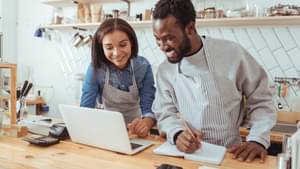 Eine Frau und ein Mann mit Schürzen schauen auf einen Laptop und informieren sich über Förderungen zu ihrem Geschäft.