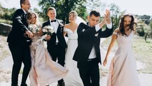 Eine junge feiernde Hochzeitsgesellschaft in Smokings und eleganten Abendkleidern, hält sich tanzend und lachend in den Armen.