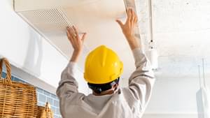 Ein Mann mit Schutzhelm bringt eine Belüftungsanlage an der Decke eines Raumes an.