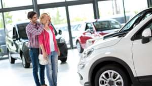 Mann und Frau stehen in einem Autohaus vor einem Auto. Er steht hinter ihr und hält ihr die Augen zu.