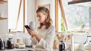 Eine Frau lehnt sich mit Kaffeetasse und Smartphone in der Hand gegen eine Arbeitsplatte in der Küche und schaut etwas kritisch auf ihr Handy.