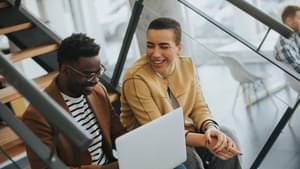 Zwei Personen sitzen lächelnd auf einer Treppe und schauen auf ein Laptop, welches der Mann auf seinen Beinen hat.