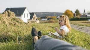 Kind sitzt in der Wiese vor moderner Wohnsiedlung neben Frauenbeinen. Aufnahme aus der Perspektive der Frau.