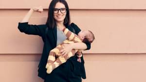 Frau mit Baby in einem Arm steht vor einer Wand und flext ihren anderen Arm. 