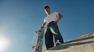 Ein Skater steht auf einer Halfpipe und schaut lässig nach unten. Dabei stützt er sich auf sein Skateboard.