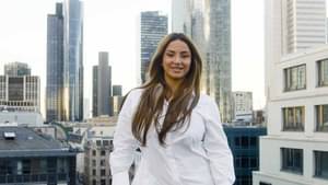 Enissa Amani steht in einem weißen Oversize-Hemd auf dem Dach eines Hauses und lächelt in die Kamera. Hinter ihr sieht man die Skyline von Frankfurt.