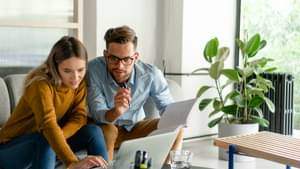 Ein Pärchen sitzt auf der Couch, schaut auf einen Laptop und macht sich dabei Notizen.