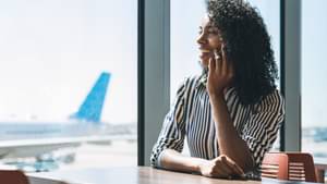 Eine Frau in einer gestreiften Bluse sitzt am Flughafen vor einem großen Fenster in der Sonne, telefoniert und lacht.