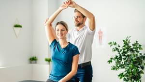 Ein Physiotherapeut macht Armbewegungsübungen mit einer jungen Frau, die auf einer Liege sitzt.