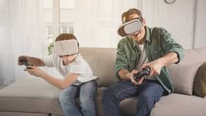 Vater und Sohn sitzen auf der Couch und spielen ein Spiel auf dem Fernseher. Beide haben einen Joystick in der Hand und tragen eine AR-Brille.