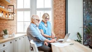 Zwei ältere Menschen stehen an einer Küchenzeile, auf dem ein Laptop steht, und informieren sich über Hinzuverdienste bei der Rente.