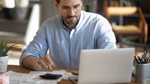 Junger Mann mit Brille und Hemd sitzt vor seinem Laptop und tippt in einen Taschenrechner.