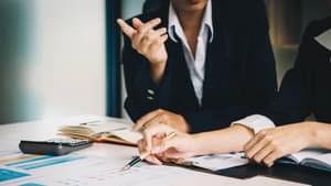 Zwei junge Frauen in schwarzen Blazern und weißen Blusen, sitzen an einem Schreibtisch und diskutieren über Bilanzergebnisse