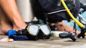 Eine Taucherbrille liegt auf einem Holzsteg vor einer sitzenden Frau mit ihrer Taucherausrüstung