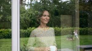 Frau steht mit ihrer Kaffeetasse am Fenster schaut ihrer Tochter beim Spielen zu.