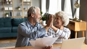 Ein älterer Mann und eine ältere Frau sitzen zusammen am Schreibtisch vor einem Laptop und geben sich lachend ein High-Five