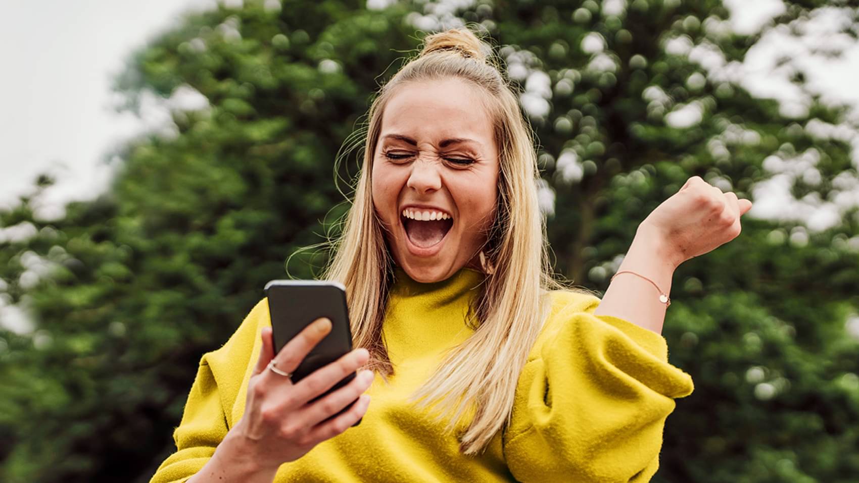 Eine junge, blonde Frau mit Zopf und gelben Pulli schaut auf ihr Handy und freut sich und hebt dabei einen Arm mit der Hand zu Faust geballt nach oben.
