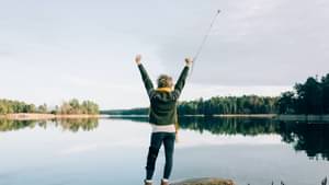 Frau mit kurzen Haaren und gelbem Schal steht mit dem Rücken zur Kamera vor einem See uns streckt beide Arme mit Krücke in der Hand nach oben.