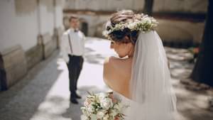 Eine junge Braut steht mit Schleier steht mit dem Rücken zur Kamera und präsentiert ihren Brautstrauß. Im Hintergrund steht ihr Ehemann