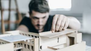 Mann im Hintergrund mit Architekturmodell im Vordergrund, an dem der Mann gerade arbeitet und sehr intensiv begutachtet.