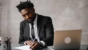 Mann im Anzug sitzt in einem modernen Büro an einem Schreibtisch. Neben ihm steht ein Laptop und er macht sich Notizen auf einem Blatt Papier.