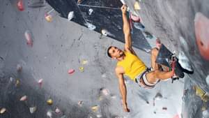 Ein Kletterer mit gelben Shirt hängt mit einer Hand an einer Boulderwand und orientiert sich nach der nächsten Griffmöglichkeit.