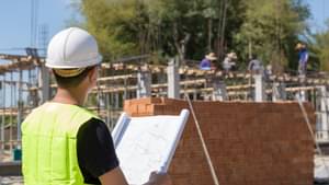 Bauleiter mit Schutzhelm und Warnweste steht mit einem Bauplan vor der Baustelle eines neuen Hauses.