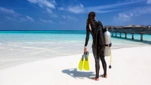 Eine sportliche junge Frau in Neoprenanzug steht mit einer Sauerstoffflasche und Schwimmflossen in der Hand an einem weißen Sandstrand und schaut auf das türkise Wasser des Meeres