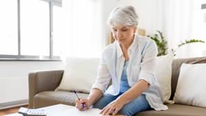 Eine ältere Frau mit weißen kurzen Haaren sitzt auf einer Couch und schaut auf einige Rechnungen. Neben ihr liegt ein Taschenrechner auf dem Couchtisch.