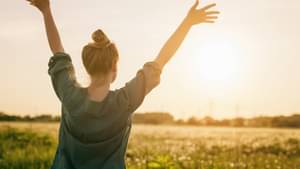 Eine junge Frau steht mit dem Rücken zur Kamera und schaut über eine grüne Wiese Richtung Sonnenuntergang und streckt beide Arme in den Himmel. 