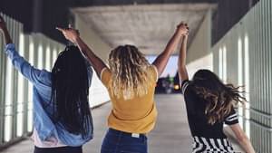 Drei junge Frauen gehen schwungvoll einen Flur entlang, halten sich an den Händen und halten dabei die Arme in die Luft.