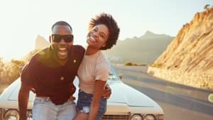 Ein junges Paar steht auf einer kurvigen Straße durch die Berge in der Sonne vor einem Auto und strahlt vor Freude.