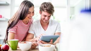 Juges Paar sitzt am gedeckten Frühstückstisch und schaut gemeinsam auf ein Tablet. Beide lächeln und er zeigt auf etwas auf dem Tablet.