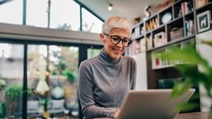 Eine Frau sitzt in einem modernen offenen Wohnzimmer und informiert sich an ihrem Laptop über Aktien.