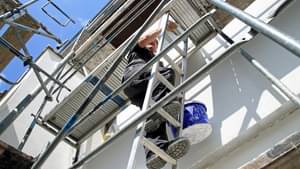 Mann mit Farbeimer in der Hand klettert ein Baugerüst hinauf.