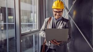 Mann im Anzug und mit gelbem Schutzhelm, steht mit einem Laptop auf dem Arm auf dem Flur eines modernen Gebäudes.