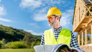 Bauingenieur mit Schutzhelm und Sicherheitsweste steht vor einem Neubau und hält den Gebäudeplan in den Händen.