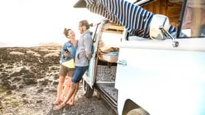 Zwei junge Reisende lehnen an einem geöffneten VW Bulli, der an einer Küste parkt.