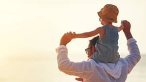 Mann steht mit Rücken zur Kamera Richtung Sonnenuntergang und hält die Arme seiner Tochter, die auf den Schultern sitzt.