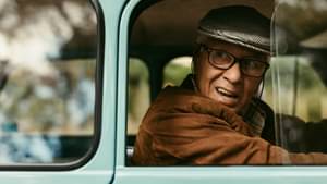 Ein älterer Herr mit Schiebermütze sitzt in einem Oldtimer und schaut über seine Schulter nach draußen.